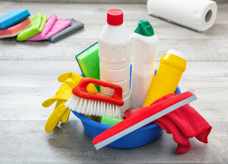 Baseboard cleaning tools including scrub brush, microfiber cloth, gloves, and cleaning products in a blue bowl on wood flooring.